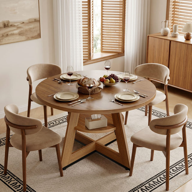 A round wooden dining table set for four, with beige upholstered chairs, is placed on a patterned rug. The table is adorned with plates, cutlery, wine glasses, a bread basket, and a bowl of fruit. The room features wooden cabinets, a painting, and large windows with blinds.